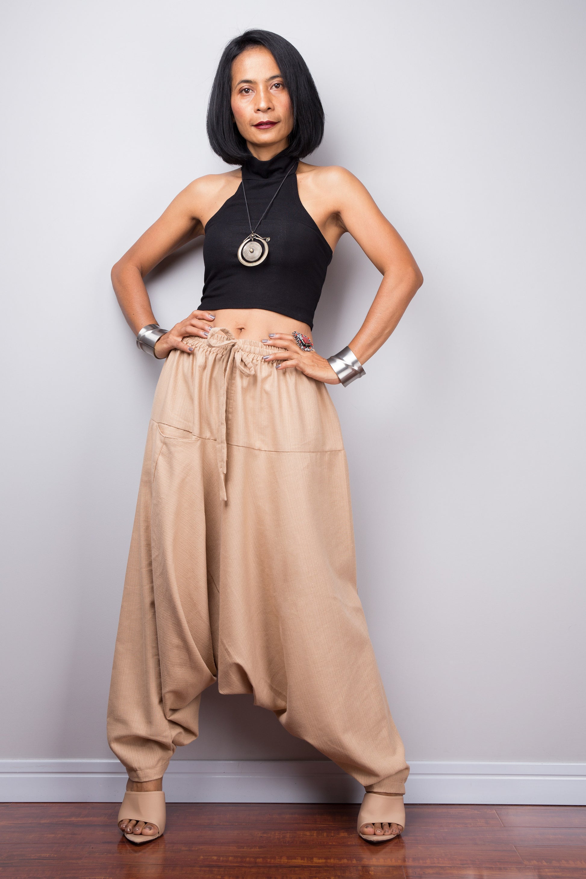 Model wearing beige harem pants with a loose fit, standing against a plain background, showcasing the full-length view. Hands positioned on hips