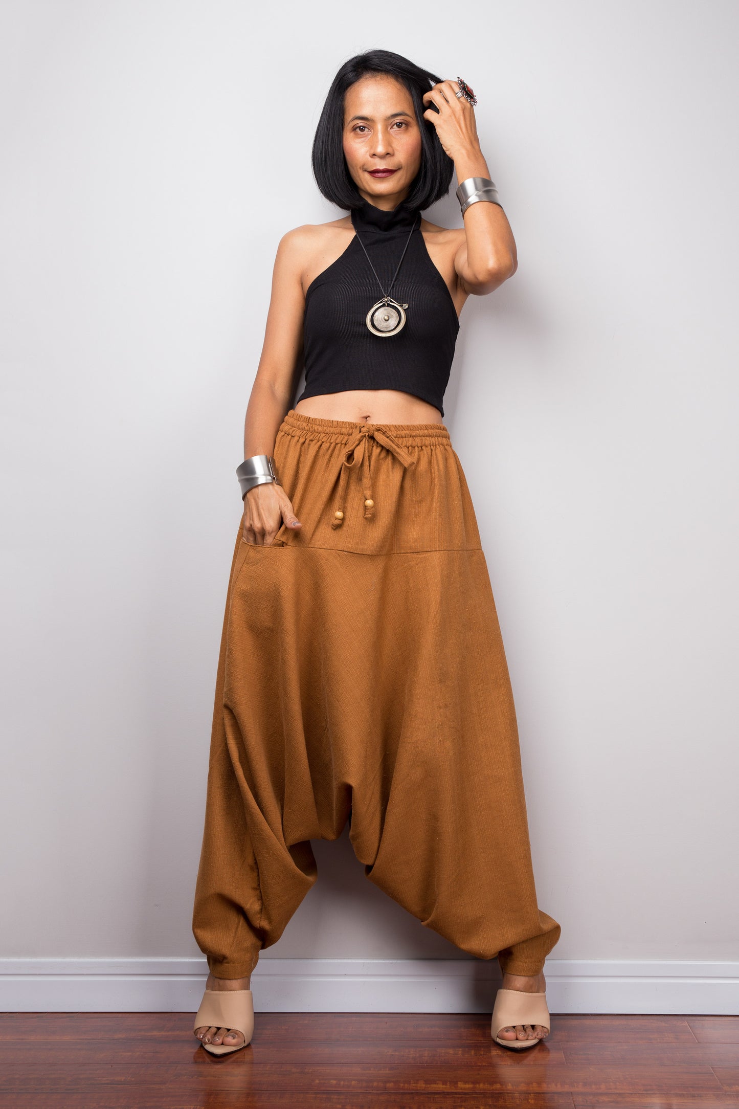 Model wearing brown harem pants with a loose fit, standing against a plain background, showcasing the full-length view. Hand in front pocket