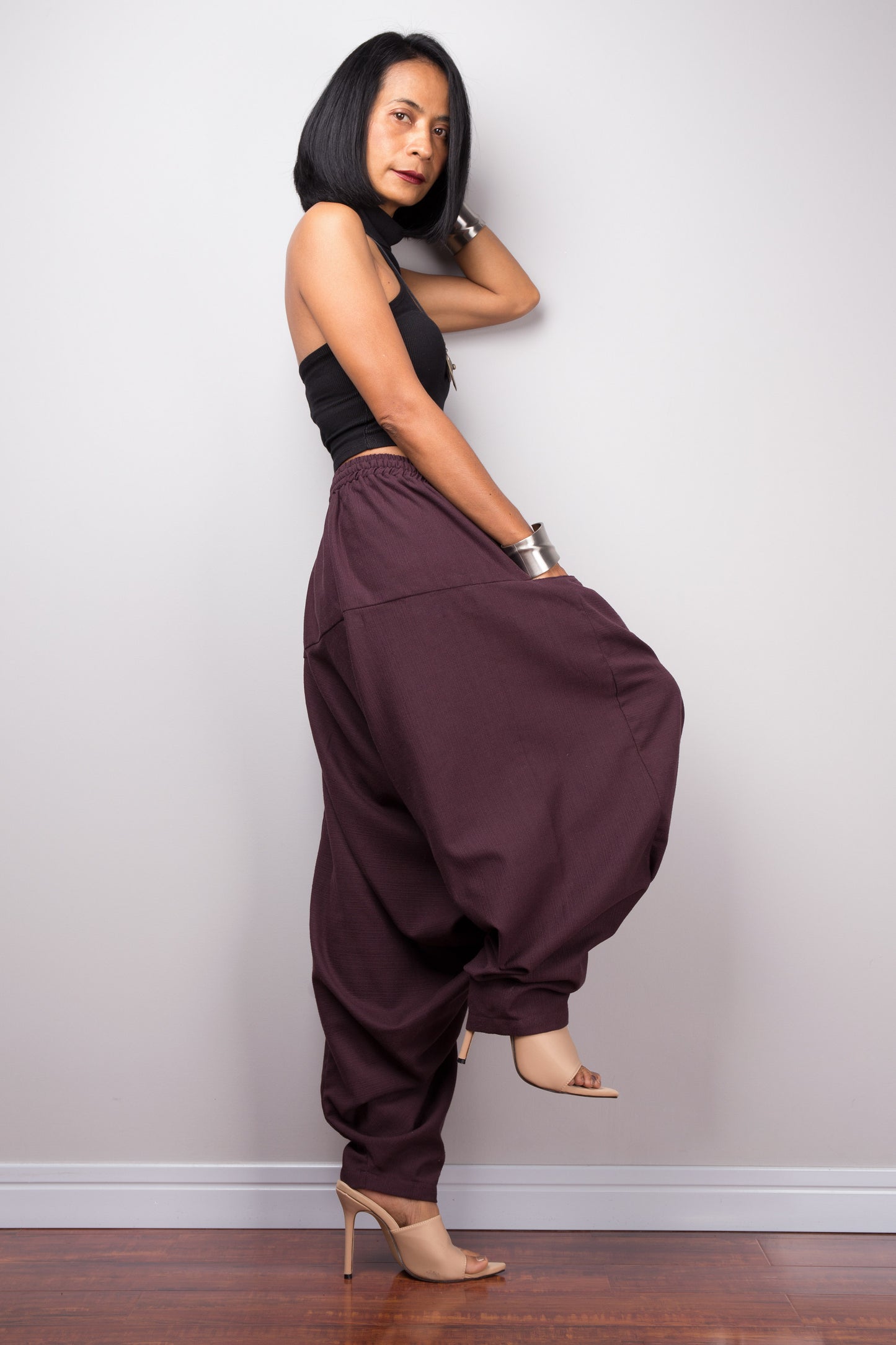 Side view of the burgundy harem pants, displaying the loose fit and the flow of the fabric from the waist to the hem. One leg raised standing pose.