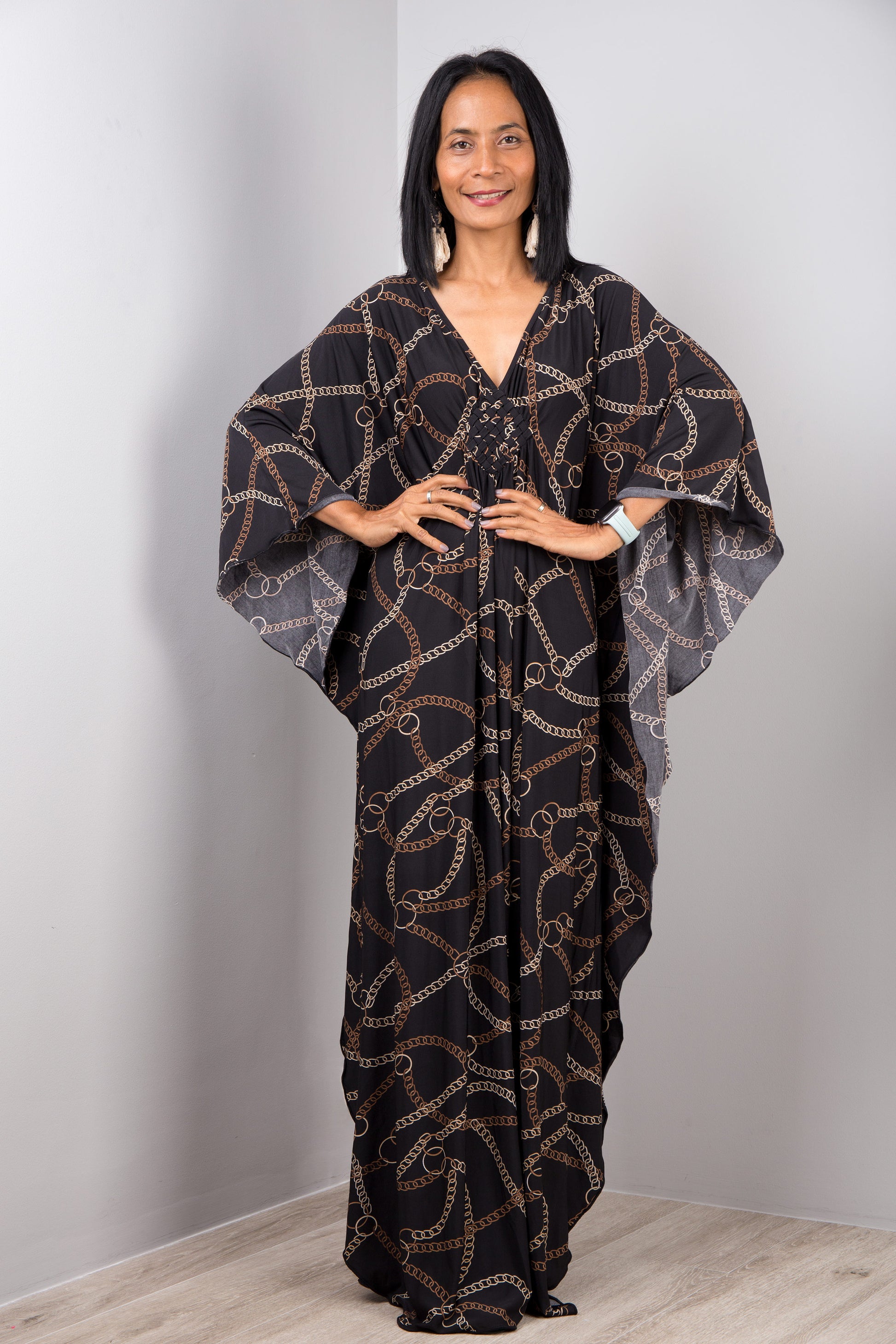 Woman in a black and gold chain-link kaftan dress by Nuichan, standing with hands on hips, showcasing the dress’s wide sleeves and maxi length, in a studio with a gray backdrop and wooden floor.