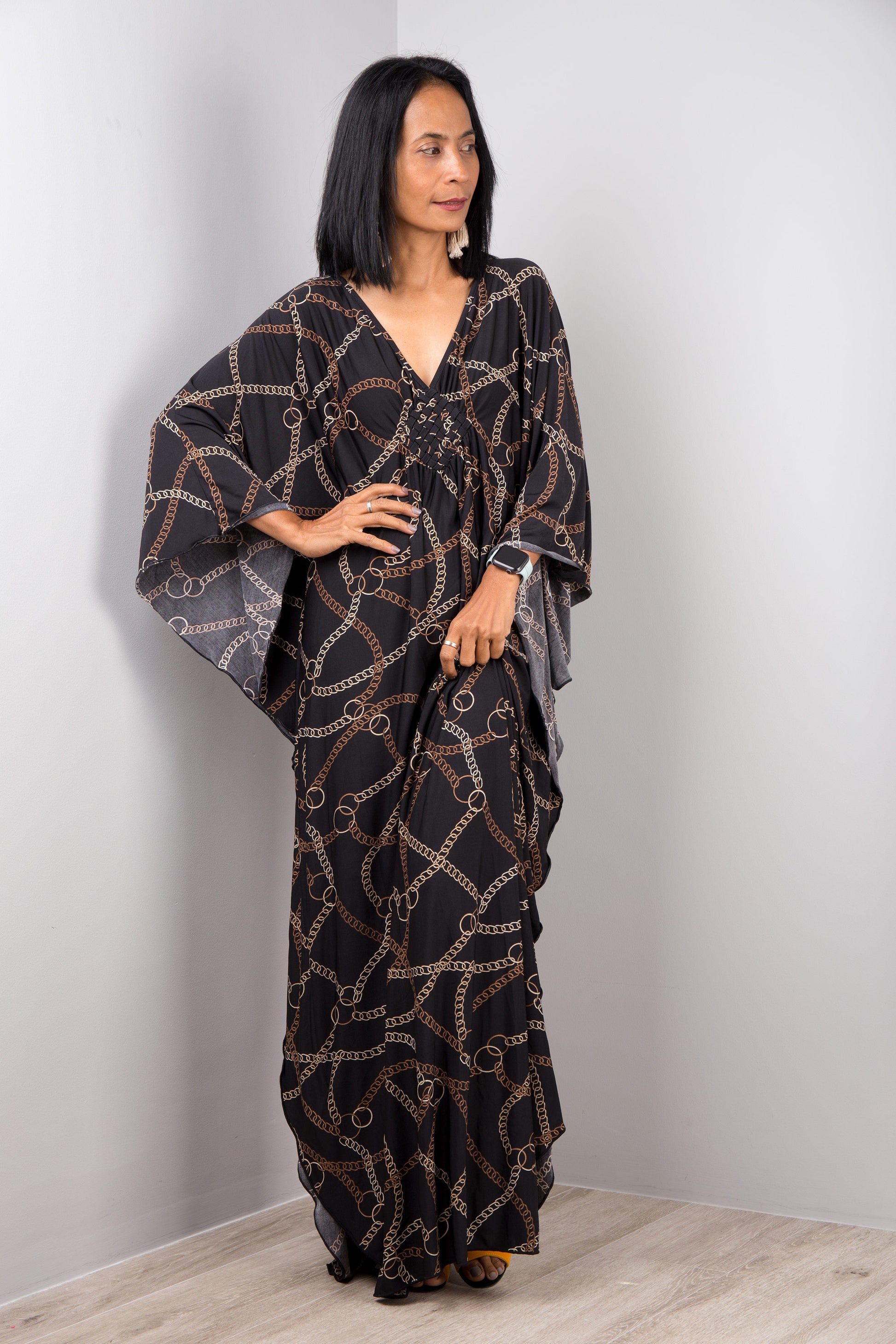 Nuichan’s black and gold chain-patterned kaftan dress, worn by a woman with dark hair and a white smartwatch, posed against a gray wall on a light wood floor, highlighting its luxurious boho style.