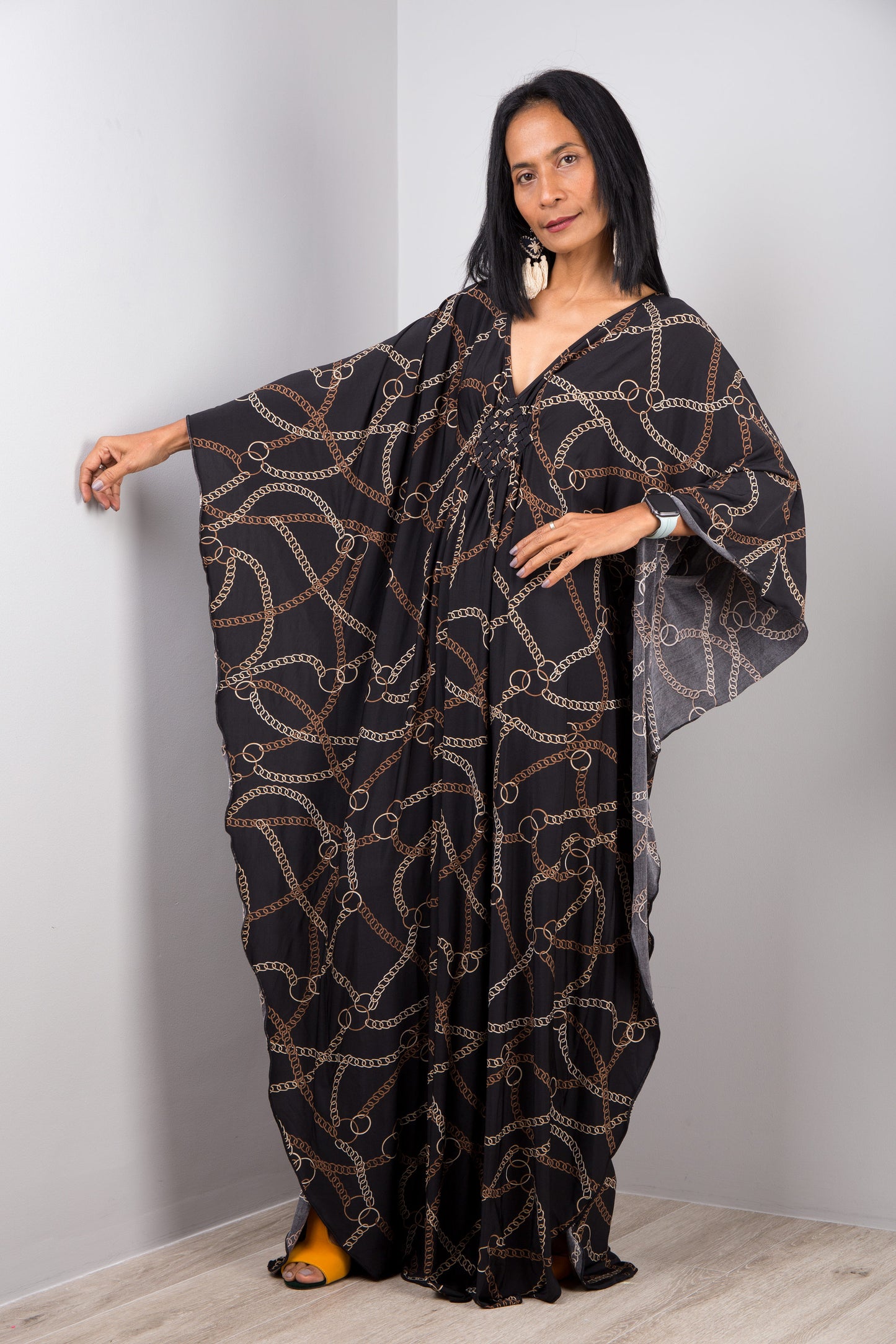 Black and gold chain-patterned kaftan dress by Nuichan, worn by a woman with long black hair, standing gracefully against a light grey wall and wooden floor, featuring wide sleeves and a floor-length design.