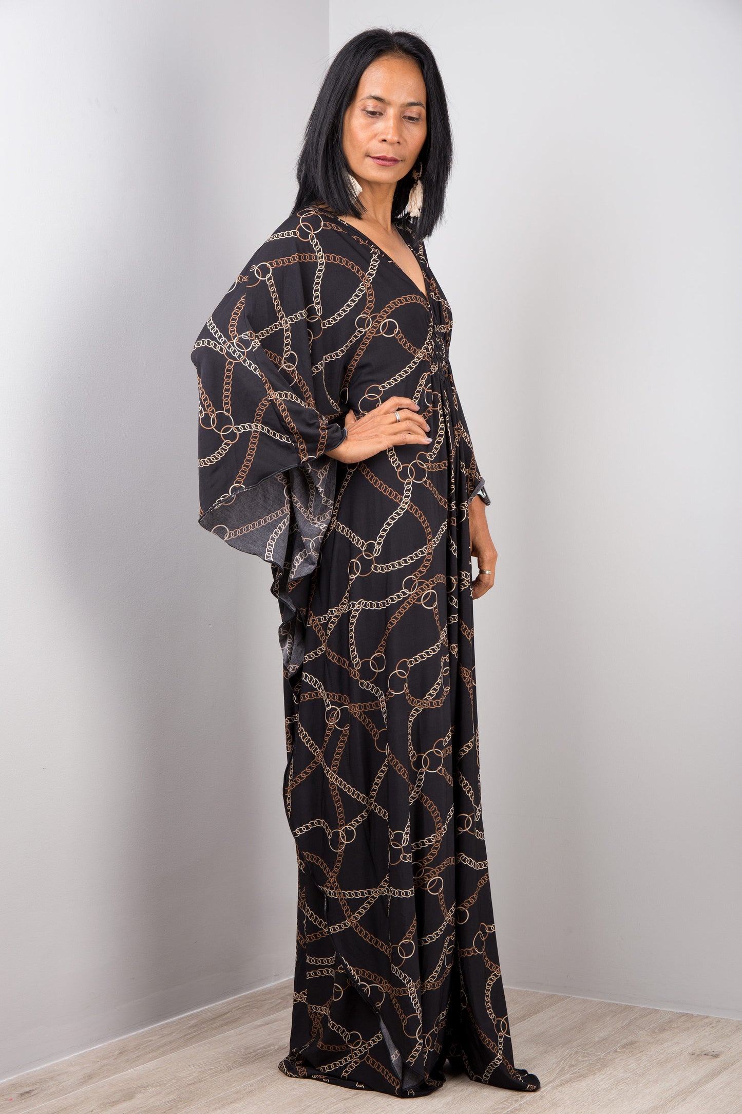 A floor-length black and gold chain-patterned kaftan dress from Nuichan, worn by a woman standing sideways, featuring loose, flowing fabric and short sleeves, set against a gray wall and wooden floor.