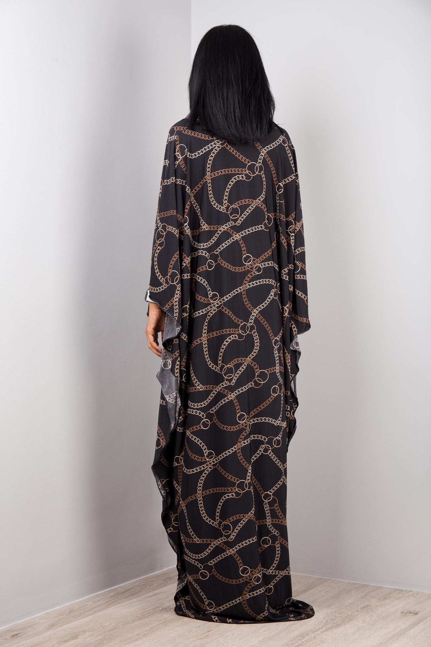 Black and gold chain-patterned kaftan dress from Nuichan, worn by a woman with dark hair and a white smartwatch, back view pose against a gray studio wall and light wood floor, highlighting its opulent boho-chic design and flowing silhouette.