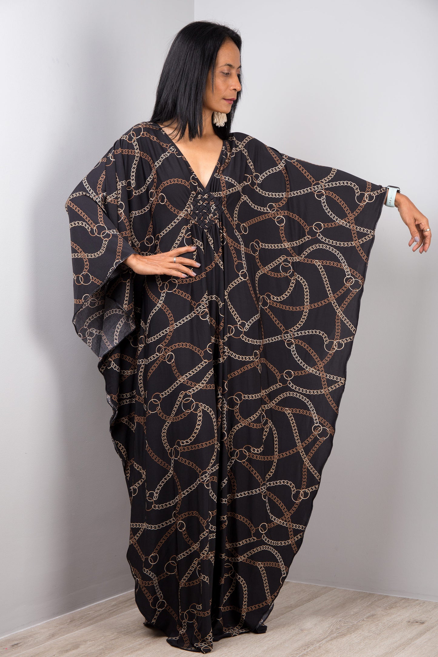 Nuichan’s black and gold chain-link kaftan dress, modeled by a woman with long black hair, posed gracefully in a minimalist studio with a gray background and light wood flooring.