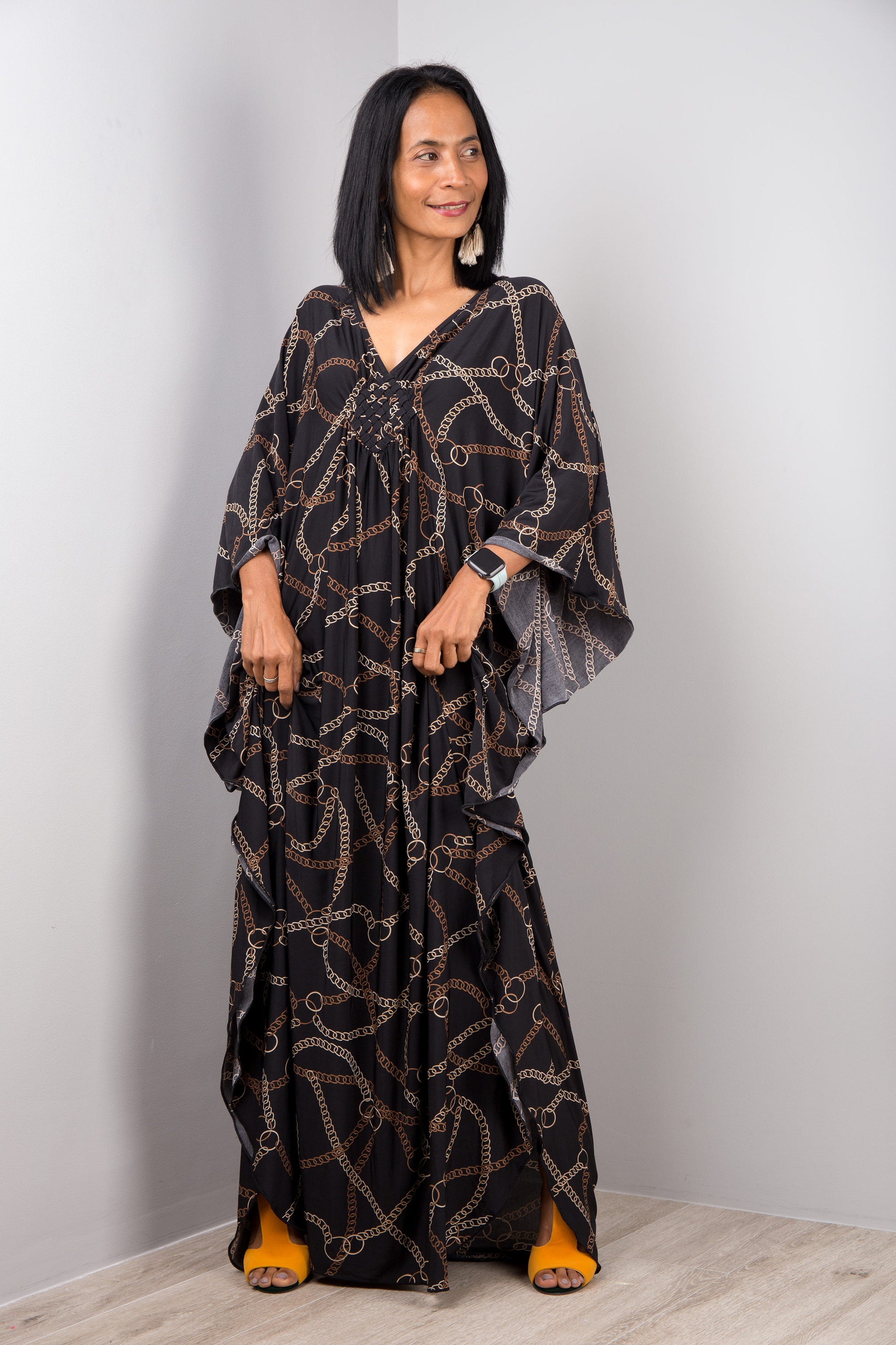 Nuichan’s floor-length kaftan dress in black and gold chain patterns, modeled by a woman in a studio with a gray background and light wood floor, wearing a white smartwatch and tassel earrings.