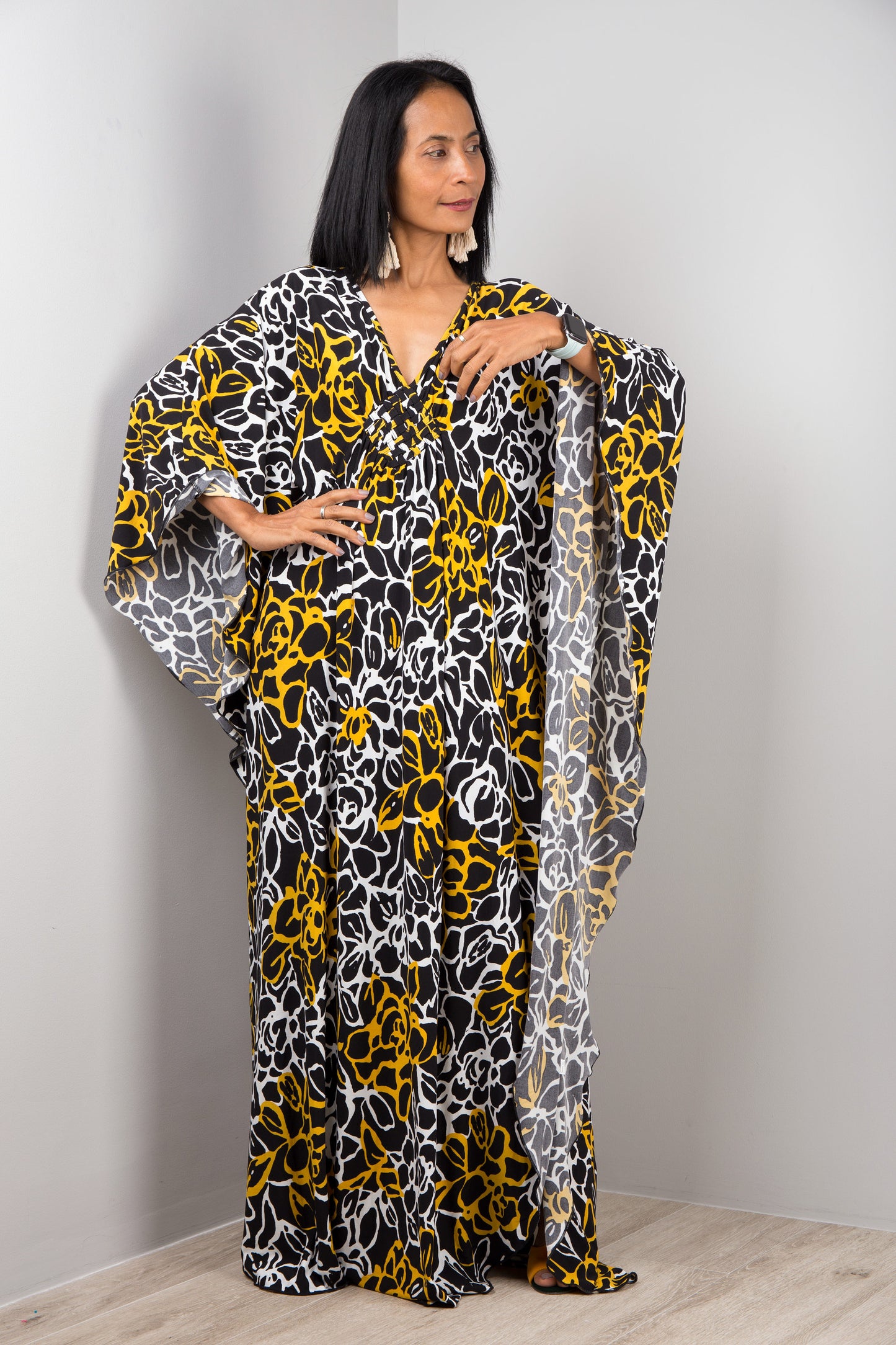 Nuichan’s black and yellow tie-dye kaftan dress with swirling floral patterns, worn by a woman with dark hair and a white smartwatch, posed against a gray wall on a light wood floor, highlighting its boho style.