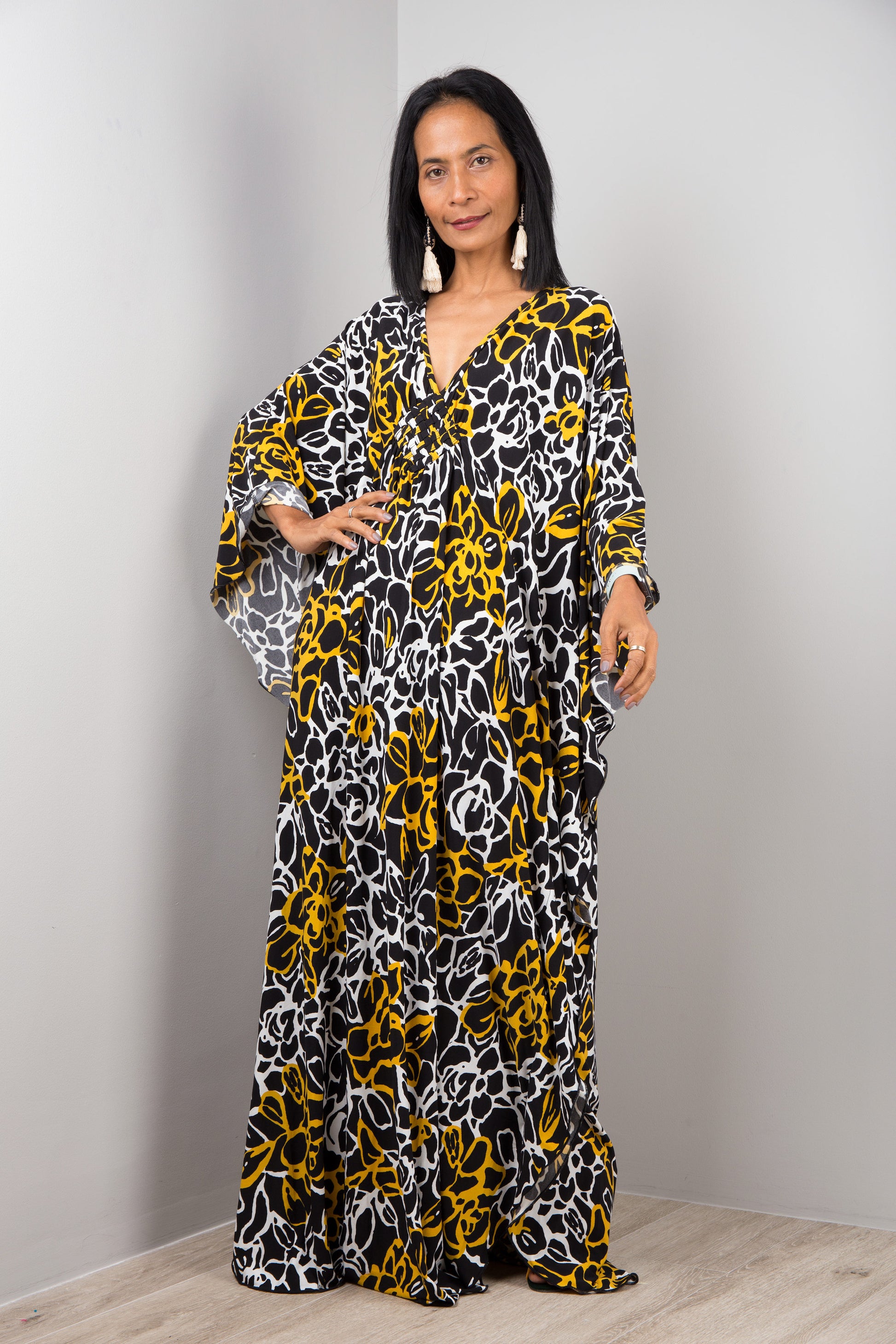 Woman in a black, yellow, and white abstract floral kaftan dress by Nuichan, standing with hands on hips, showcasing the dress’s wide sleeves and maxi length, in a studio with a gray backdrop and wooden floor.