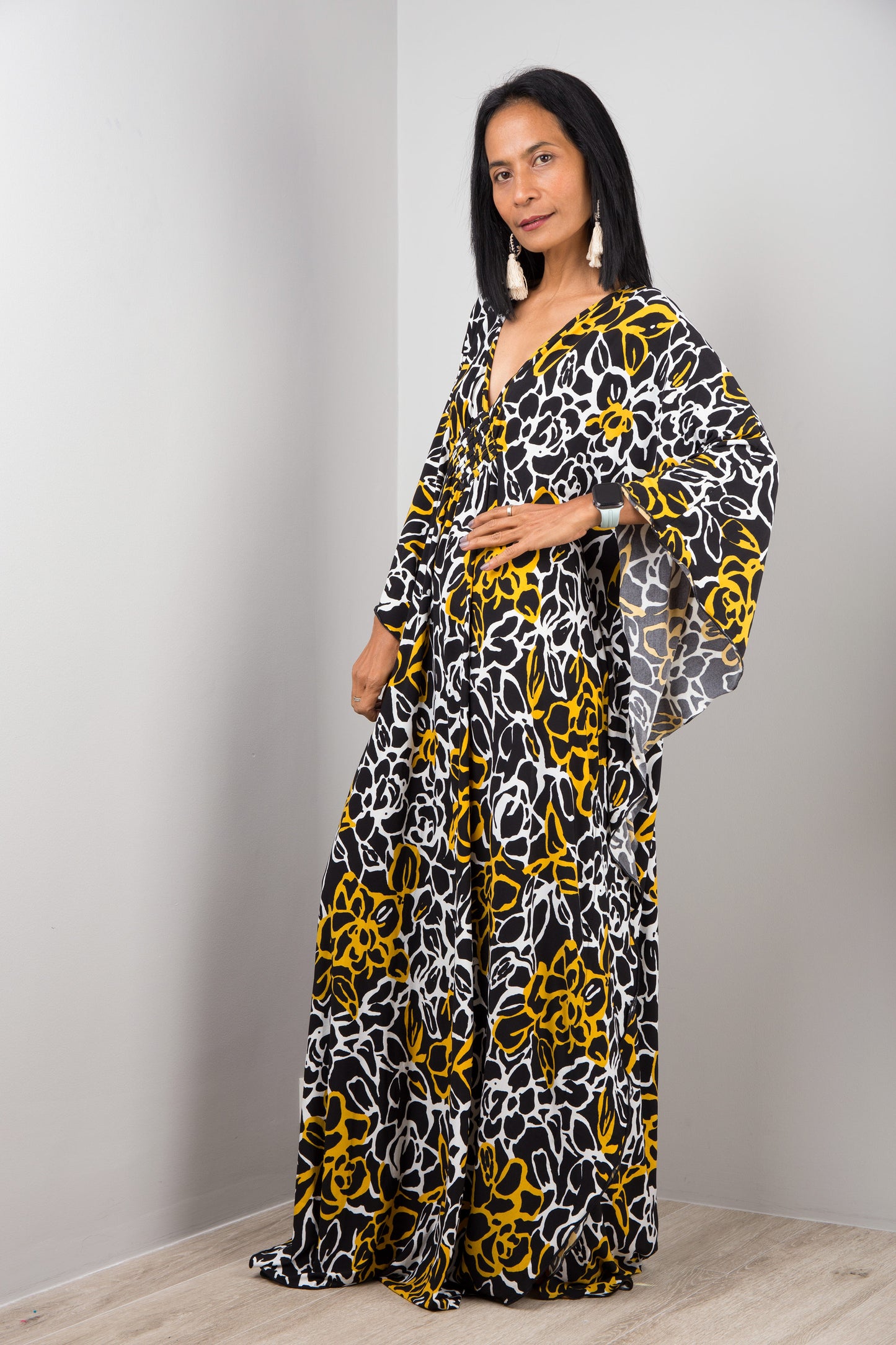 A floor-length black, white, and yellow floral kaftan dress from Nuichan, worn by a woman standing sideways, featuring loose, flowing fabric and short sleeves, set against a gray wall and wooden floor.