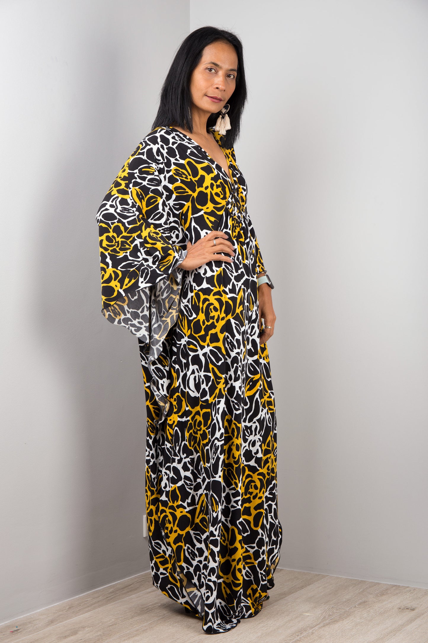 A woman wearing Nuichan’s black and yellow floral kaftan dress with white accents, standing sideways in a minimalist studio, showcasing the dress’s loose fit, wide sleeves, and maxi length against a gray wall and wooden floor.