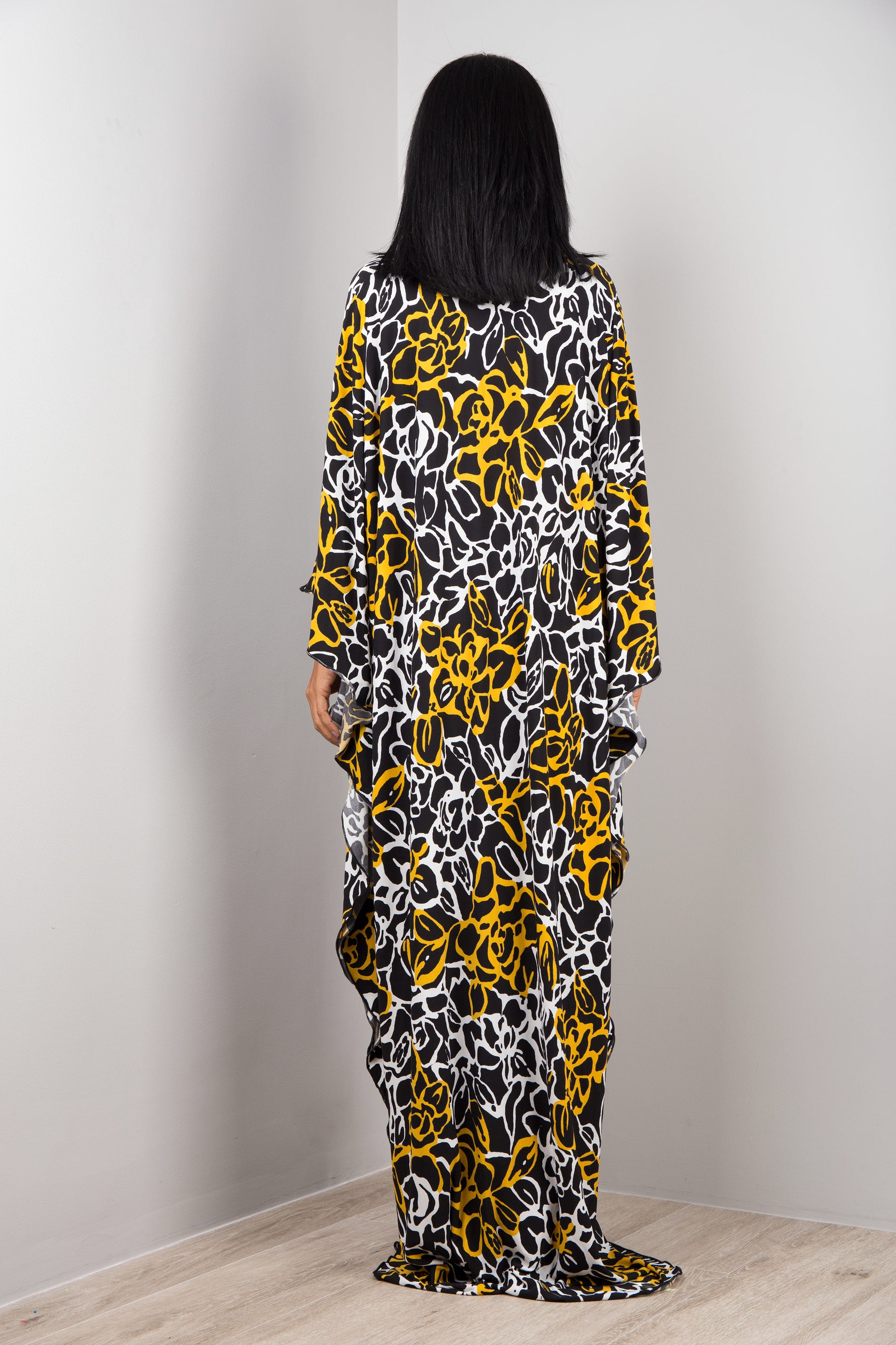 Black, white, and yellow tie-dye floral kaftan dress from Nuichan, worn by a woman with dark hair and a white smartwatch, posed against a gray studio wall and light wood floor, highlighting its boho-chic design and flowing silhouette.