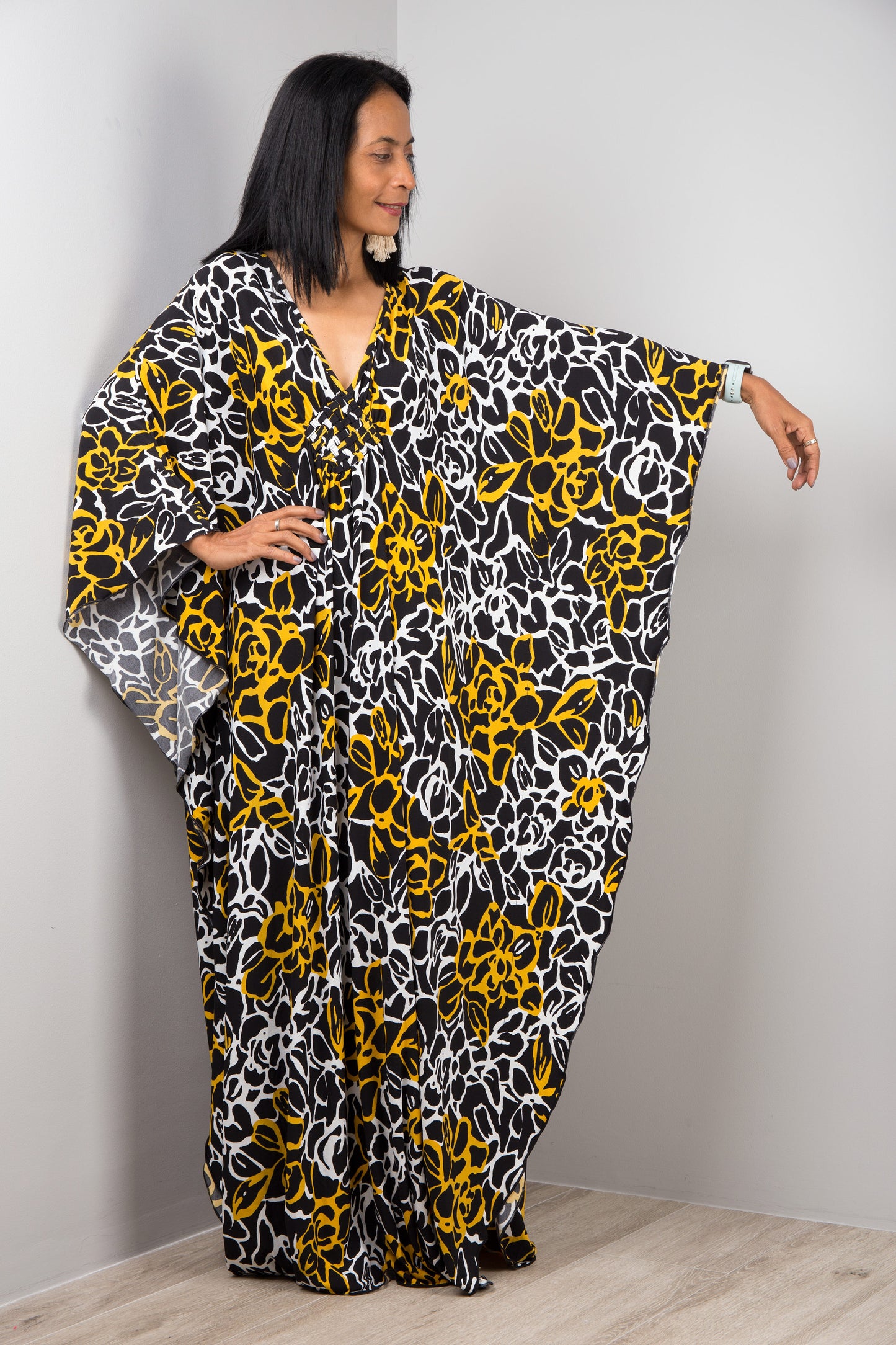 A woman wearing a black, yellow, and white floral-patterned kaftan dress, standing against a plain gray wall on a wooden floor, with wide, flowing sleeves and a V-neckline, accessorized with a white smartwatch and tassel earrings.