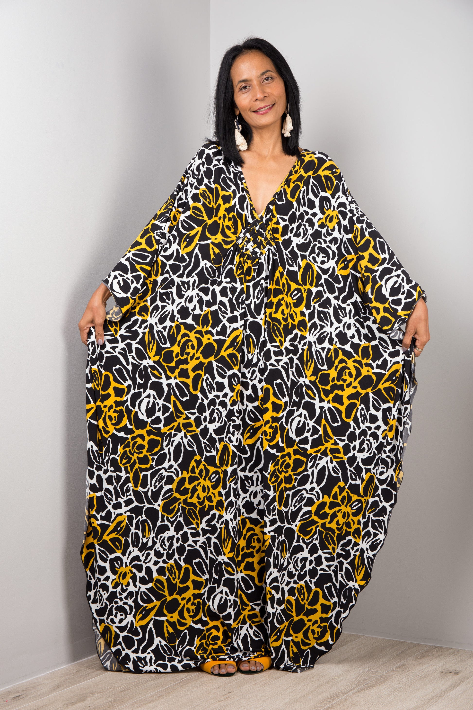 A woman modeling a black, white, and yellow floral-patterned kaftan dress from Nuichan, with a loose fit, V-neck, and flowing sleeves, standing in a simple gray and wood studio setting, accessorized with earrings.