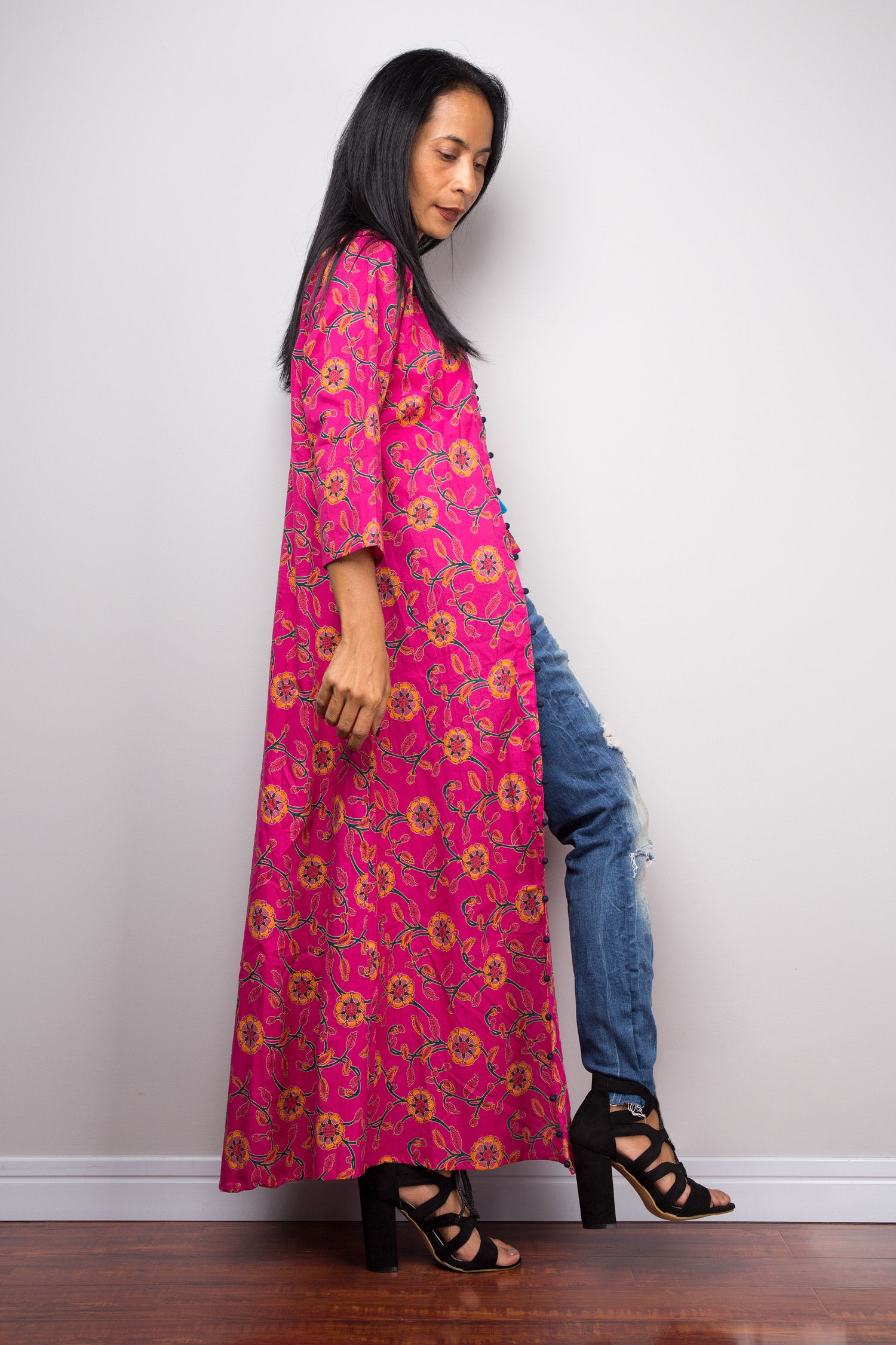 Back view of a woman in a pink floral cotton cardigan, standing against a grey wall with her hand in her hair.