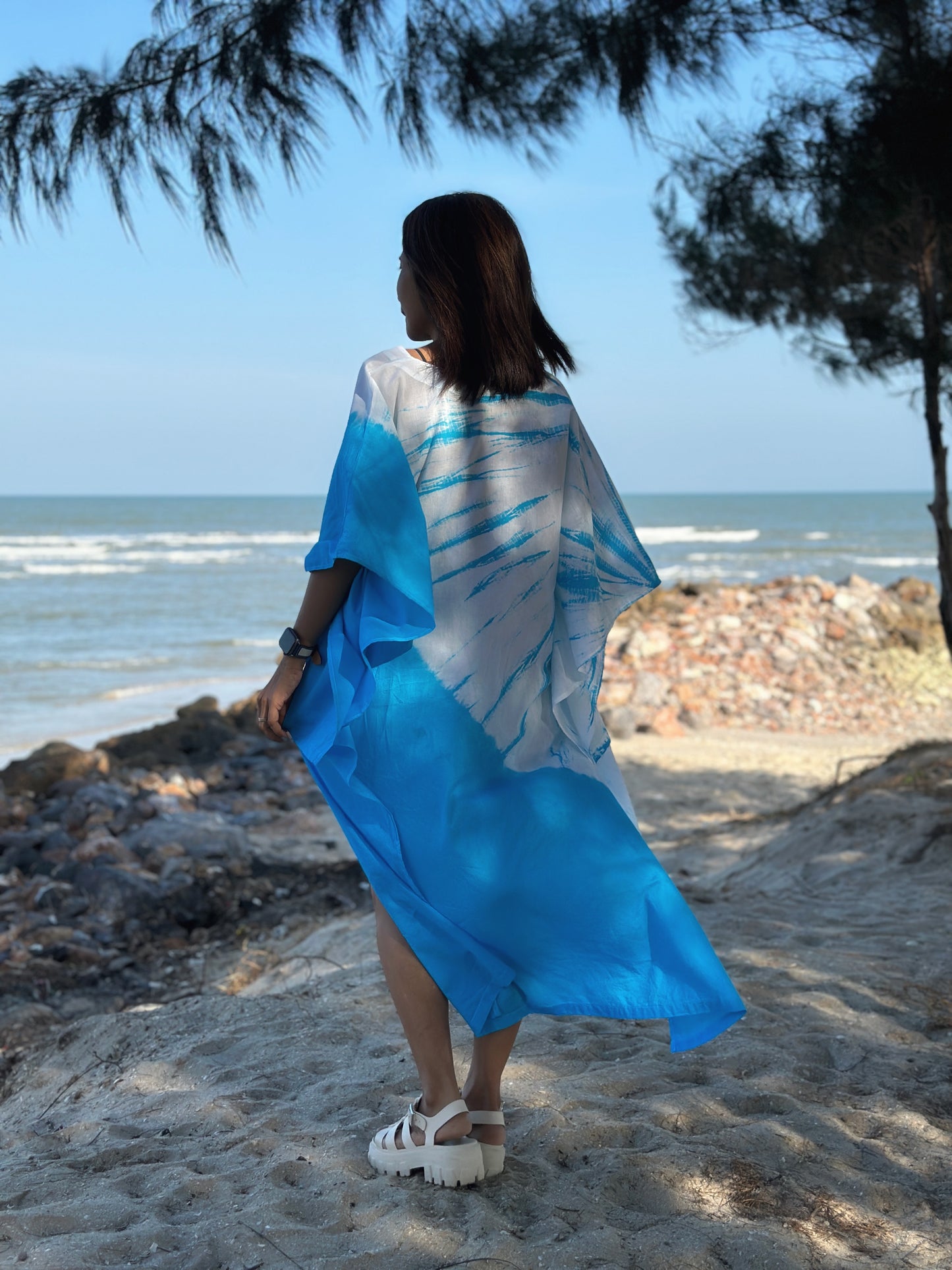 Back view of a woman wearing a light cotton short front kaftan dress with blue shibori tie dye pattern, standing by the beach with ocean waves in the background.