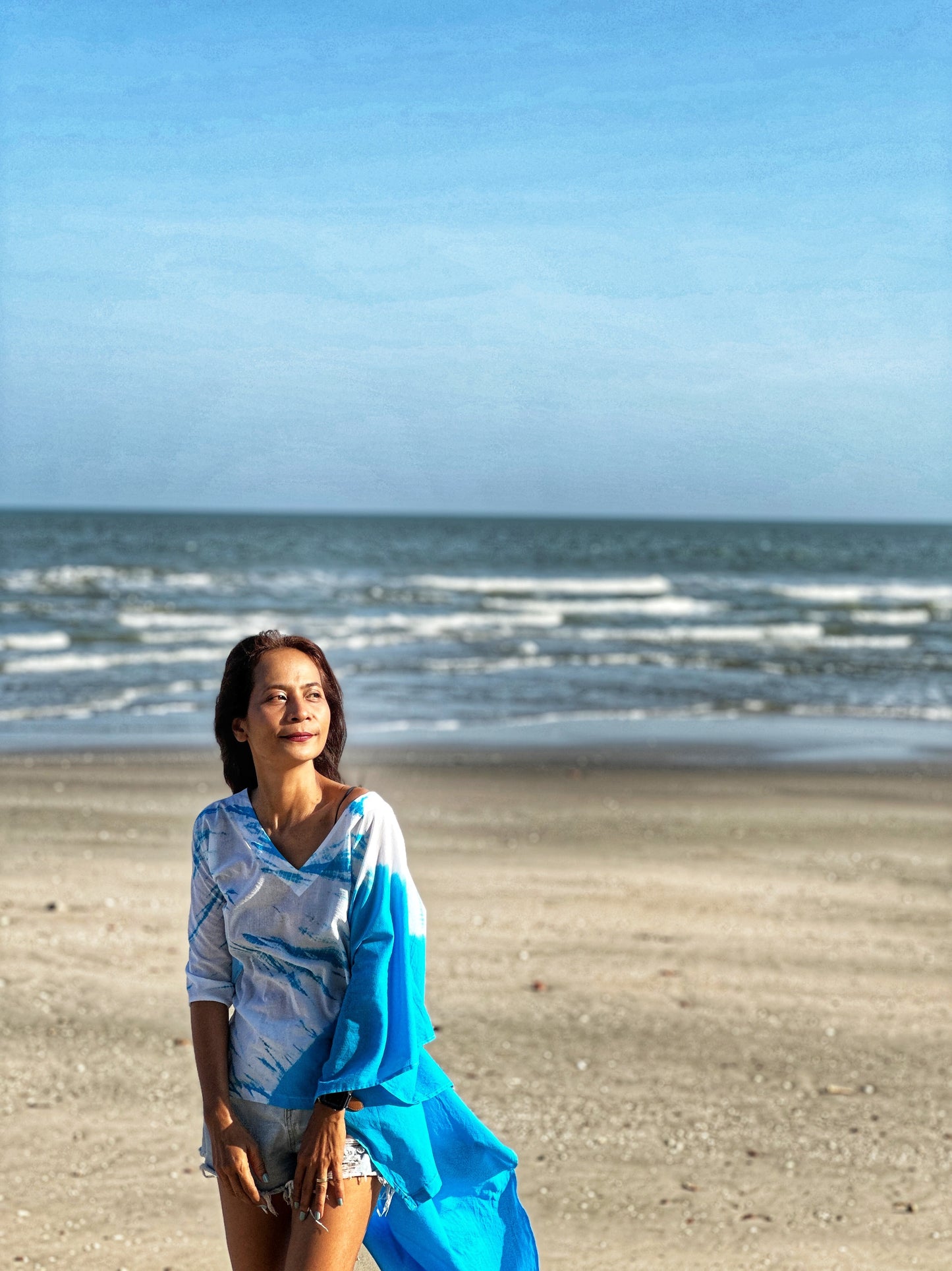 "Fashionable short front kaftan dress in blue shibori tie dye, worn casually over shorts, with the wearer walking on a sandy beach