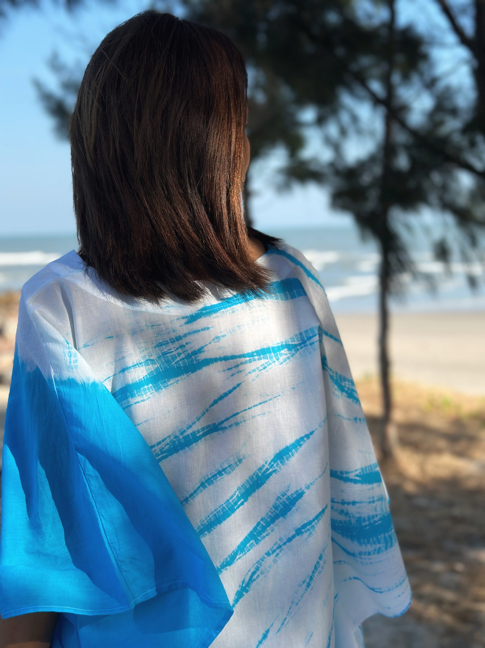 Close-up of the intricate blue shibori tie dye pattern on the back of a short front cotton kaftan dress, worn by a woman on a beach.