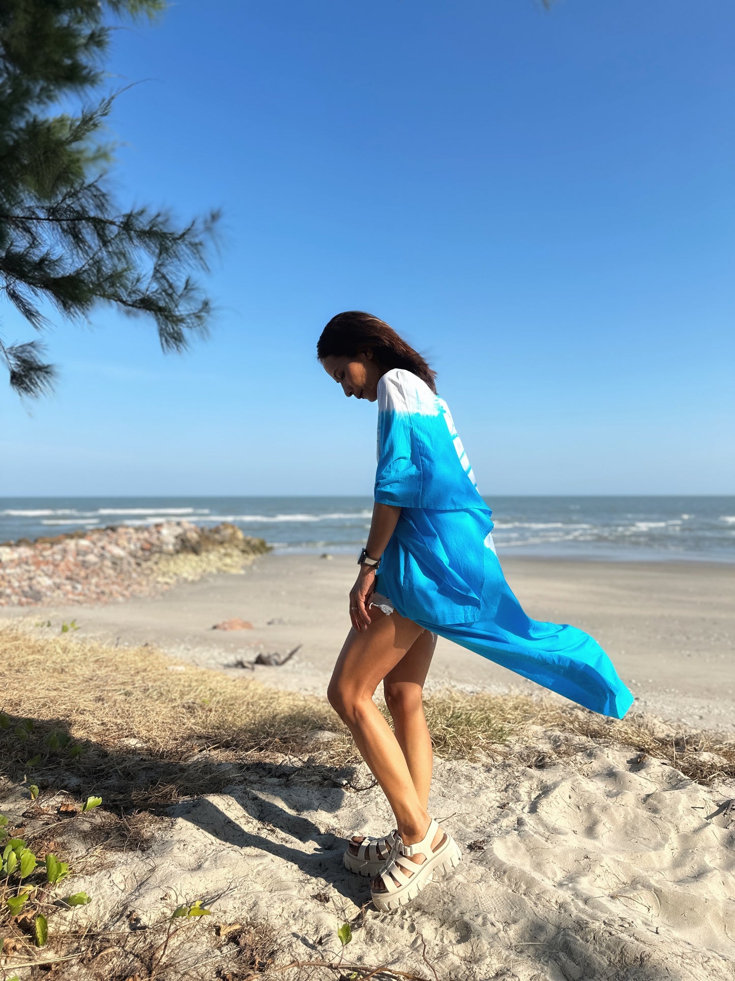 Side profile of a woman in a blue shibori tie dye kaftan dress walking on the beach, with the ocean and rocky shore in the distance.