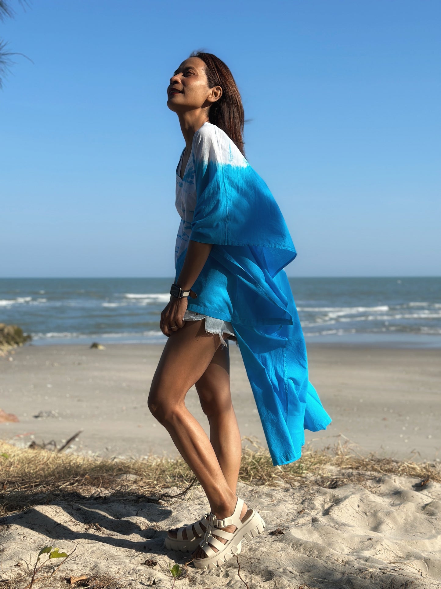 Beachside elegance with a woman in a short front kaftan dress, the blue tie dye fabric flowing as she walks, ocean waves in the background.