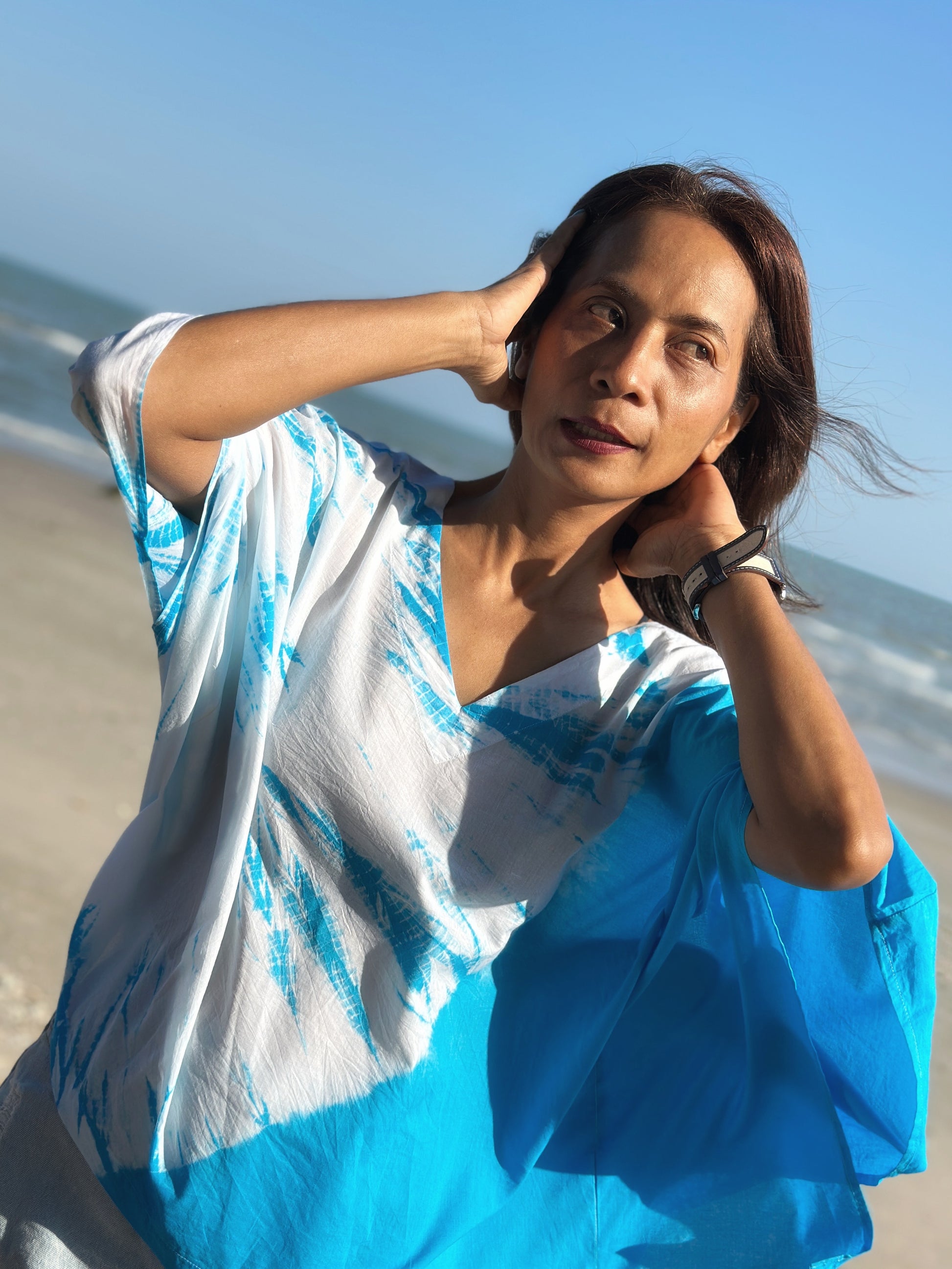 A woman poses on the beach in a light cotton kaftan dress with a striking blue and white tie dye pattern, highlighting the short front style.