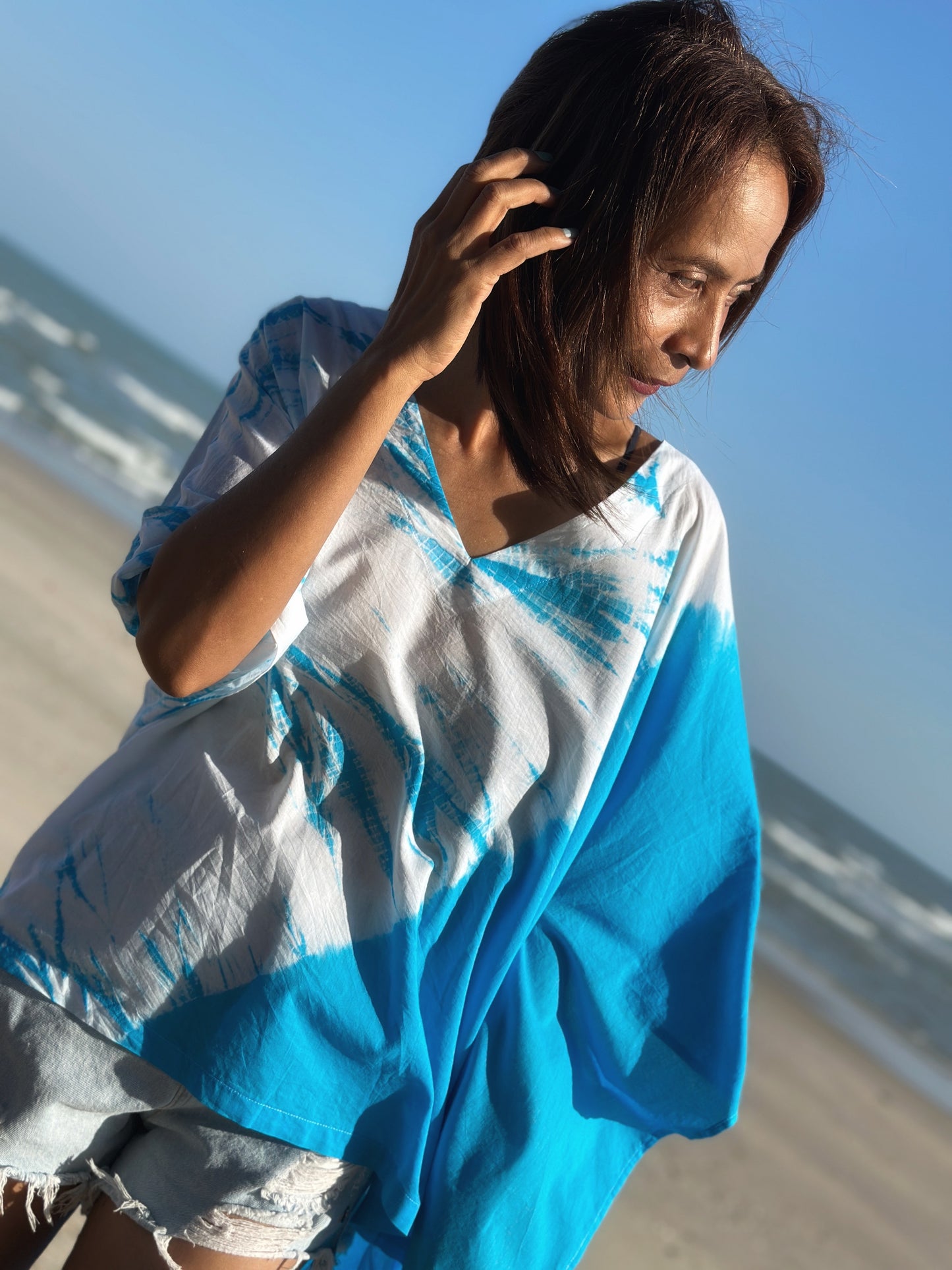 Vibrant blue and white shibori tie dye kaftan dress worn over denim shorts, captured in a beach setting under a clear sky.