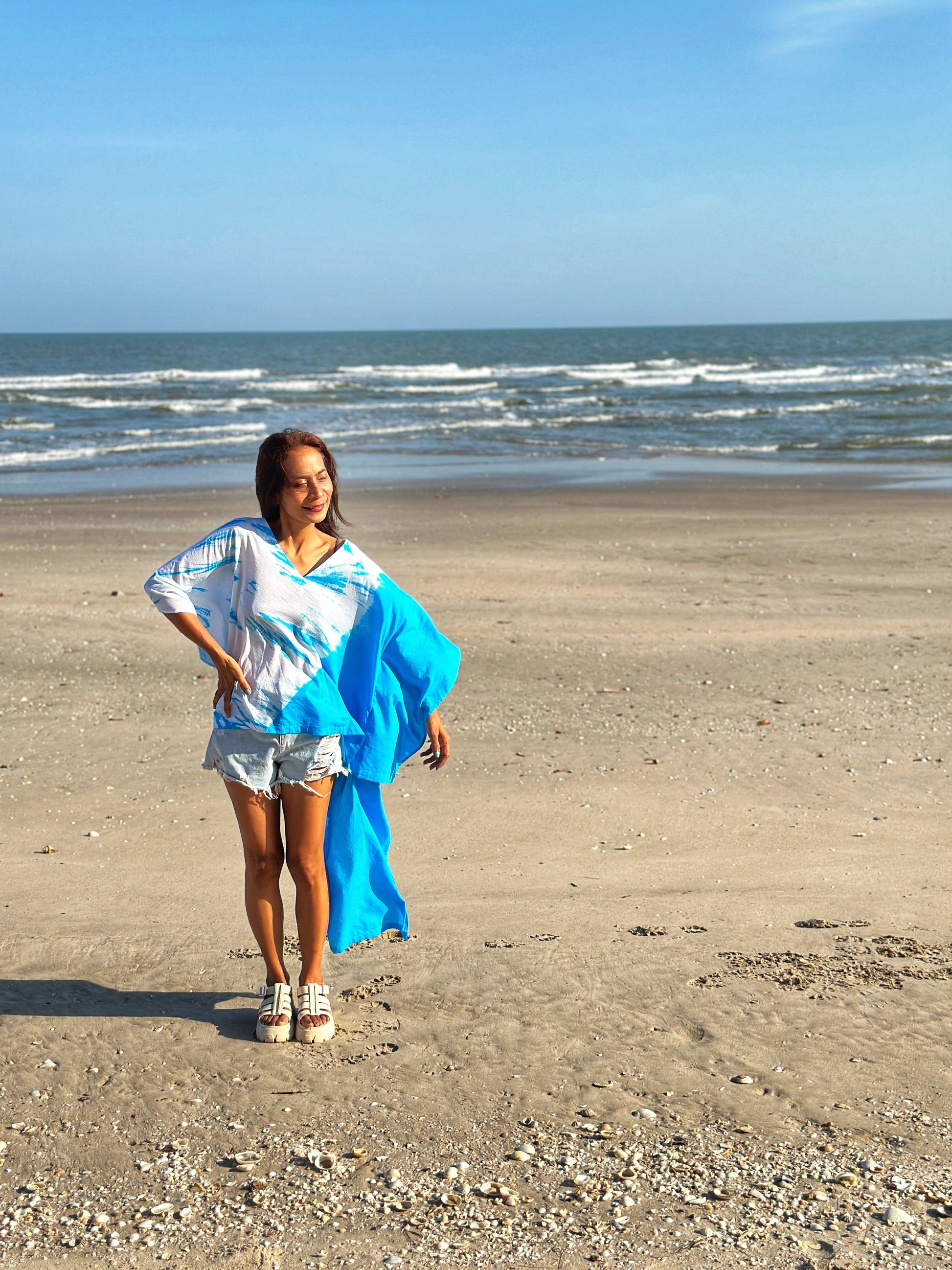 A woman strolls along the beach wearing a light cotton kaftan dress with vibrant blue tie dye, showcasing the short front design.